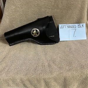 Op de afbeelding: Zwarte leren holster met een zilveren en gouden starembleem. De holster is gestikt met witte draad en is gelabeld "LEFT HANDED BLK".
