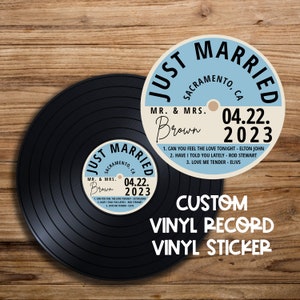 Op de afbeelding: Een blauwe en witte vinyl platen sticker met de tekst "Just Married" en "Sacramento, CA" in zwart. De sticker bevat ook de datum "04.22.2023" en de namen "Mr. & Mrs. Brown". De sticker zit op een zwarte vinyl plaat.