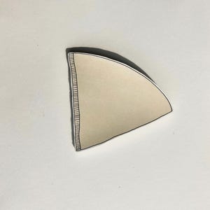 May include: A beige, cone-shaped coffee filter made of paper. The filter features a distinct, reinforced vertical seam along one straight edge, characterized by a series of fine, dark, parallel lines that provide structural integrity to the paper.