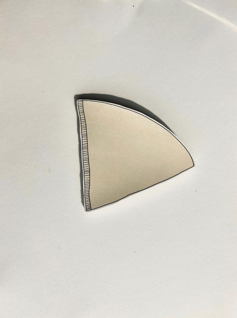 May include: A beige, cone-shaped coffee filter made of paper. The filter features a distinct, reinforced vertical seam along one straight edge, characterized by a series of fine, dark, parallel lines that provide structural integrity to the paper.