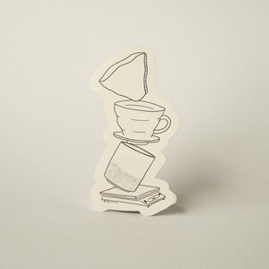 Può includere: Un disegno in bianco e nero di un'attrezzatura per la preparazione del caffè. Un filtro per il caffè è posto sopra un filtro per il caffè in ceramica, che è posto sopra una caraffa di vetro. La caraffa è appoggiata su una bilancia.