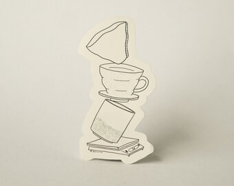 May include: A black and white line drawing of a coffee brewing setup. A coffee filter sits on top of a ceramic pour-over coffee dripper, which is placed on top of a glass carafe. The carafe is sitting on a scale.