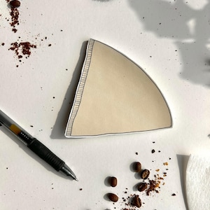Puede incluir: Un filtro de café de papel beige con forma triangular, un contorno negro dibujado a mano y un borde estilo costura en un lado. El filtro está colocado sobre una superficie blanca junto a un bolígrafo negro y granos de café tostados.
