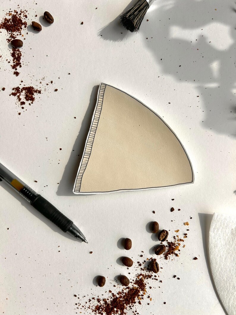 May include: A beige, triangular paper coffee filter with a black hand-drawn outline and a stitched-style border along one side. The filter is positioned on a white surface alongside a black ballpoint pen and scattered roasted coffee beans.