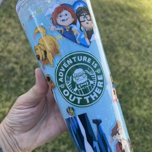 May include: A blue and green reusable tumbler with a glitter finish. The tumbler features various cartoon characters and the text "Adventure is out there".