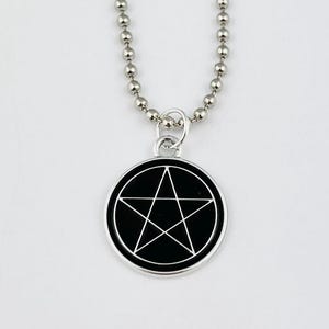 Peut inclure: Un collier à chaîne à billes argenté avec un pendentif circulaire. Le pendentif présente un fond en émail noir avec un motif d'étoile pentagramme argenté fin, centré dans un bord en métal argenté en relief et un petit anneau de jonction.