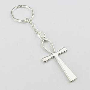 以下が含まれることがあります： アンクのシンボルペンダントが付いたシルバー調の金属製キーホルダー。アンクは短い金属チェーンで標準的な円形の二重リングに取り付けられています。全体的に滑らかで光沢のある仕上げで、シンプルでクリーンなデザインです。
