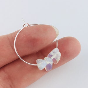 May include: A silver hoop earring featuring three iridescent, rough-cut stones. The stones are milky white and lavender. The earring is held by a person's fingers against a white background.