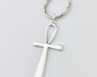 Pode incluir: Um porta-chaves de metal prateado com um pendente em forma de ankh. O ankh tem uma superfície lisa e polida e está preso a uma argola circular texturizada por um pequeno elo de ligação. O design é simples, metálico e refletor.