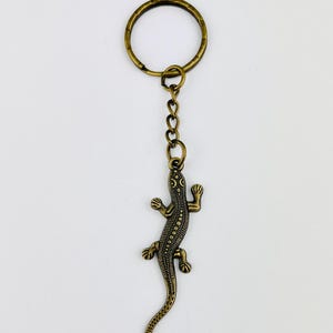 May include: A bronze-toned metal keyring featuring a lizard charm suspended from a circular ring by a short chain. The lizard charm has a textured surface with a dotted pattern along its back and four legs extending from its body.