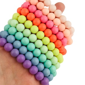 Op de afbeelding: Een verzameling kleurrijke ronde kralen in een regenboog van tinten, waaronder paars, blauw, groen, geel, oranje, roze en perzik. De kralen worden in een hand gehouden en tonen het levendige kleurenpalet.