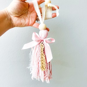 May include: A handmade tassel ornament in shades of pink and cream. The tassel is made of pink yarn with a pink satin ribbon and a wooden charm that says "Blessed". The ornament is suspended by a cream-colored ribbon and wooden beads.