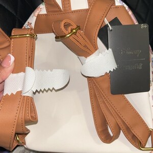 Peut inclure: Sac &agrave; dos blanc et beige avec des bretelles marron et des ferrures dor&eacute;es. Le sac &agrave; dos pr&eacute;sente un motif floral sur les c&ocirc;t&eacute;s et une &eacute;tiquette noire avec l'inscription "Disney Loungefly". Les bretelles sont r&eacute;glables.