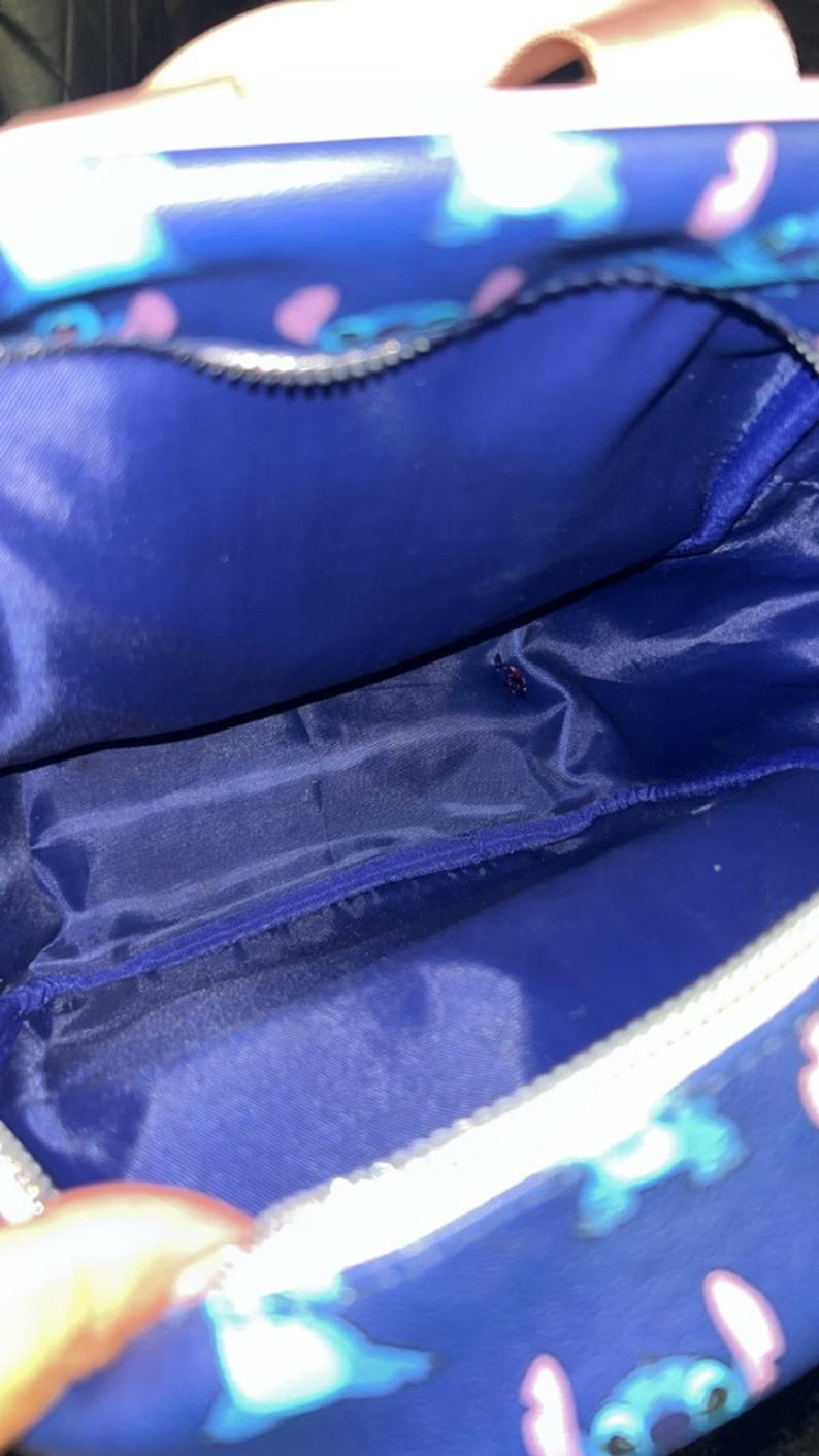 May include: A blue cosmetic bag with a white zip and a Stitch character pattern. The interior is a solid blue colour. The bag is designed to hold makeup and other beauty products.