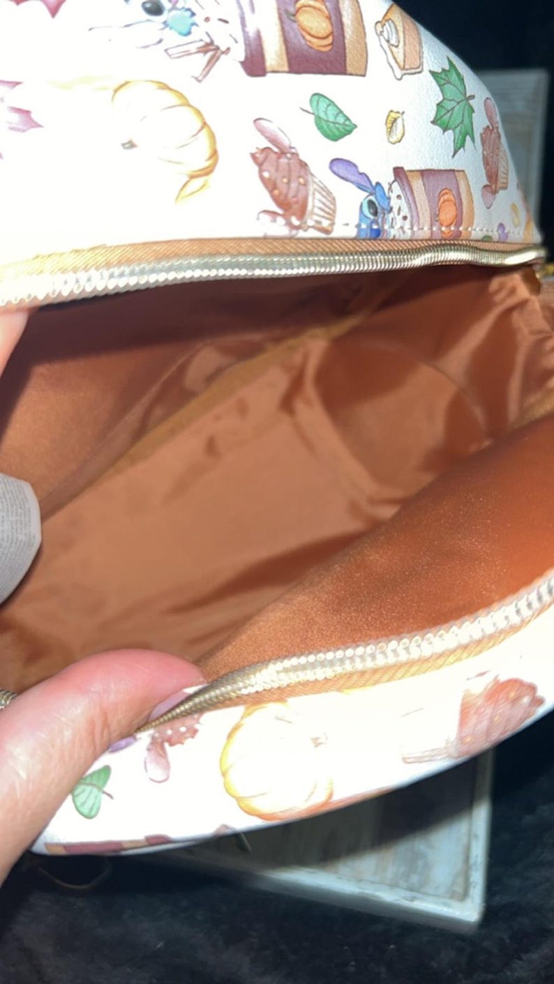 Peut inclure: Sac cosm&eacute;tique blanc ouvert avec fermeture &eacute;clair dor&eacute;e. L'ext&eacute;rieur pr&eacute;sente un motif r&eacute;p&eacute;t&eacute; de personnages de dessins anim&eacute;s, de citrouilles et d'aliments d'automne. L'int&eacute;rieur est de couleur marron uni.