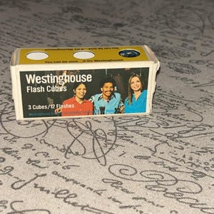 May include: A vintage Westinghouse Flash Cubes box. The box is rectangular with a yellow top and white sides. The front features the Westinghouse logo and the text "Flash Cubes". It also shows a picture of three people. The box contains 3 cubes for 12 flashes.
