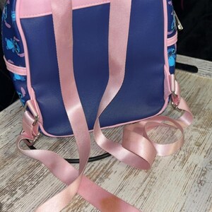 May include: A small, navy blue backpack with pink detailing and straps. The backpack features a cartoon character design and a zip closure. The pink straps are adjustable and have a satin finish. The bag is on a white wooden tray.