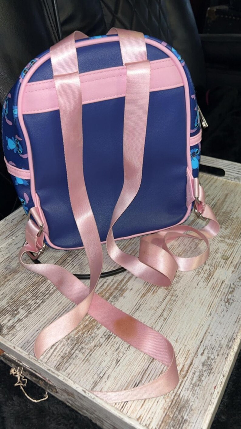 May include: A small, navy blue backpack with pink detailing and straps. The backpack features a cartoon character design and a zip closure. The pink straps are adjustable and have a satin finish. The bag is on a white wooden tray.