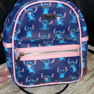 May include: A navy blue backpack with a repeating pattern of the character Stitch. The backpack has pink trim, straps, and a top handle. A front pocket with a silver zip is also present. The word "Stitch" is visible on the front.
