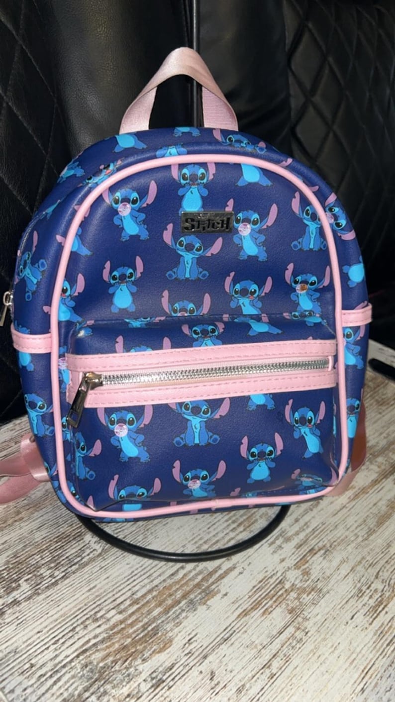 May include: A navy blue backpack with a repeating pattern of the character Stitch. The backpack has pink trim, straps, and a top handle. A front pocket with a silver zip is also present. The word "Stitch" is visible on the front.