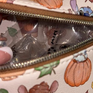 Peut inclure: Un sac cr&egrave;me avec une fermeture &eacute;clair marron, orn&eacute; d'un motif de cupcakes, de citrouilles, de feuilles et d'un personnage extraterrestre bleu. L'int&eacute;rieur du sac est visible, contenant des articles emball&eacute;s. Le mot "Made" est partiellement visible.