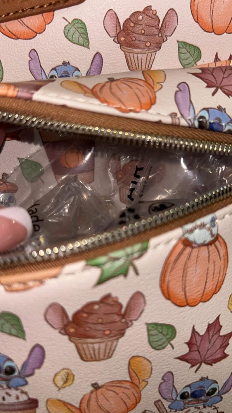 Peut inclure: Un sac cr&egrave;me avec une fermeture &eacute;clair marron, orn&eacute; d'un motif de cupcakes, de citrouilles, de feuilles et d'un personnage extraterrestre bleu. L'int&eacute;rieur du sac est visible, contenant des articles emball&eacute;s. Le mot "Made" est partiellement visible.