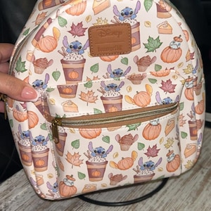 Peut inclure: Mini sac &agrave; dos blanc avec un motif automnal : citrouilles, feuilles et personnages Stitch. Il poss&egrave;de une poign&eacute;e et une fermeture &eacute;clair marron, ainsi qu'une petite poche avant. Les logos Disney et Loungefly sont visibles.