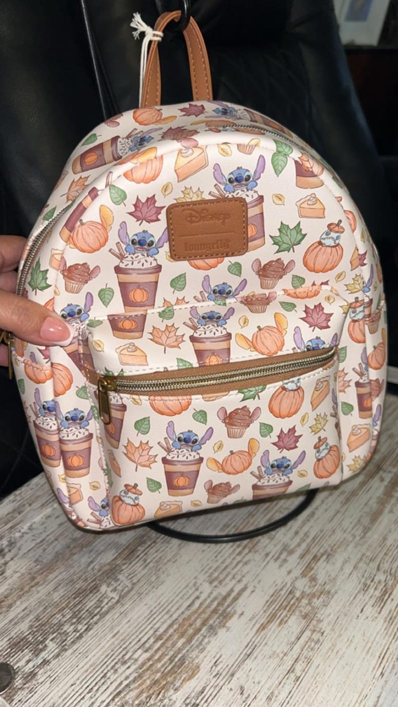 Peut inclure: Mini sac &agrave; dos blanc avec un motif automnal : citrouilles, feuilles et personnages Stitch. Il poss&egrave;de une poign&eacute;e et une fermeture &eacute;clair marron, ainsi qu'une petite poche avant. Les logos Disney et Loungefly sont visibles.