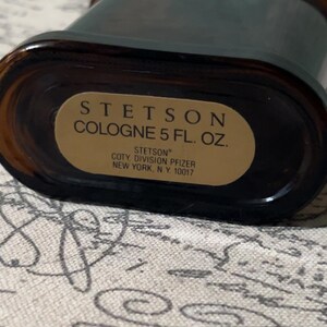 Może przedstawiać: Brązowa, prostokątna butelka wody kolońskiej Stetson ze złotą etykietą. Na etykiecie widnieje napis "STETSON COLOGNE 5 FL. OZ." i informacje producenta. Butelka ma ciemnobrązowy odcień i zaokrągloną g&oacute;rę.