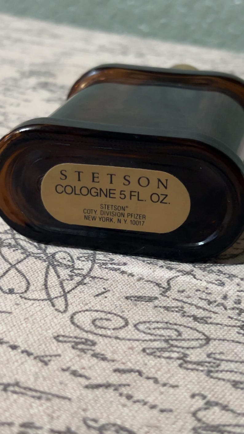 Może przedstawiać: Brązowa, prostokątna butelka wody kolońskiej Stetson ze złotą etykietą. Na etykiecie widnieje napis "STETSON COLOGNE 5 FL. OZ." i informacje producenta. Butelka ma ciemnobrązowy odcień i zaokrągloną g&oacute;rę.