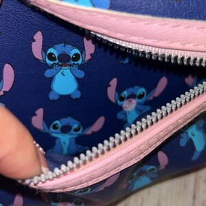 May include: A navy blue bag with a pink zip and strap, featuring a repeating pattern of the Disney character Stitch in various poses. The interior lining is white. The bag's design includes Stitch's blue fur, large eyes, and distinctive ears.