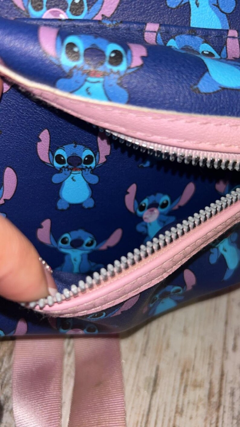 May include: A navy blue bag with a pink zip and strap, featuring a repeating pattern of the Disney character Stitch in various poses. The interior lining is white. The bag's design includes Stitch's blue fur, large eyes, and distinctive ears.
