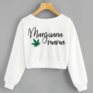 Può includere: Felpa bianca a maniche lunghe e taglio corto con un motivo a foglia di marijuana verde. Il testo "Marijuana mama" è stampato in nero sul retro della felpa.