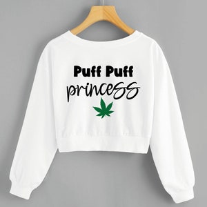 Peut inclure: Sweat-shirt blanc court à manches longues. Le devant présente le texte "Puff Puff princess" en noir, avec un graphisme de feuille de cannabis verte en dessous. Le sweat-shirt est suspendu à un cintre en bois.