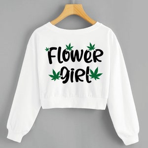 May include: White cropped sweatshirt with long sleeves and a black graphic print that says "Flower girl" with green marijuana leaf accents.