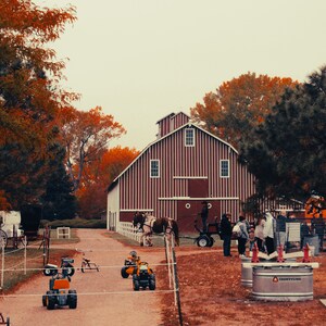 May include: A rural scene featuring a red and white barn with vertical stripes, surrounded by trees with autumn foliage. A dirt path leads to the barn, with small vehicles and a horse-drawn carriage visible. People are gathered near the barn.