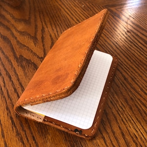 May include: A brown leather notebook cover with a stitched edge. The cover is open to reveal a blank, gridded notebook.