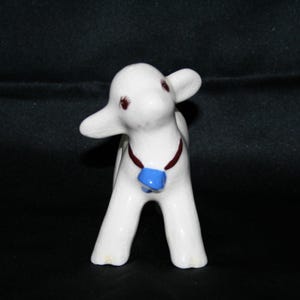 May include: A white ceramic figurine of a lamb with brown painted eyes and a blue and brown painted collar with a bell.