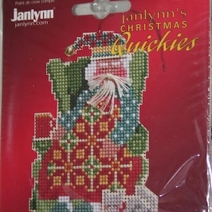 May include: A counted cross stitch pattern featuring a whimsical Christmas gnome wearing a red, green, and blue patchwork outfit. The pattern is titled "Janlynn's Christmas Quickies" and includes the phrase "Back Home" in French and English.