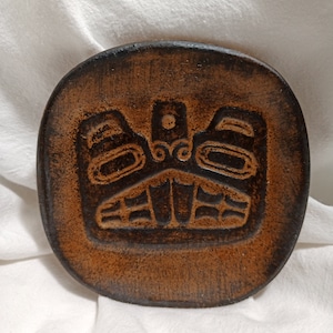 Peut inclure: Un dessous de verre rond et brun avec un motif d'oiseau stylisé en relief au centre. Le dessous de verre a une bordure marron foncé et une surface texturée. Le design rappelle l'art amérindien.