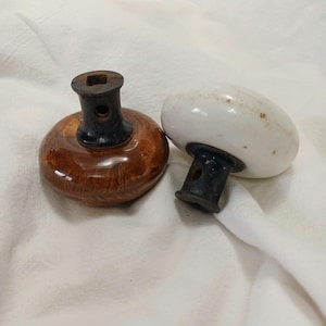 May include: Two ceramic insulators with black metal bases. One insulator is brown with a wood grain pattern, and the other is white.