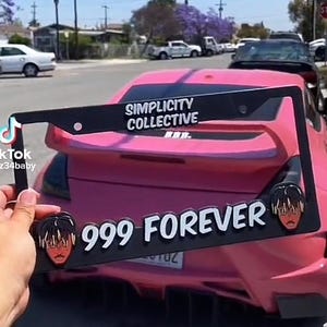 May include: A black license plate frame with the text "Simplicity Collective" and "999 Forever" in white lettering. The frame has two cartoon images of a person with black hair and a red shirt. The frame is attached to a pink car.