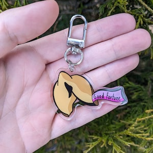 May include: A gold-colored fortune cookie keychain with a pink banner that reads "Good Fortune". The keychain has a silver metal clasp.