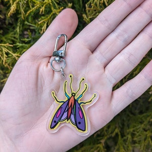 May include: A clear acrylic keychain with a colorful butterfly design. The butterfly has purple wings, a yellow body, and orange and blue accents.
