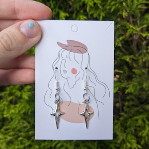 May include: A pair of silver star-shaped earrings with a simple design. The earrings are hanging from a white card with a line drawing of a woman.