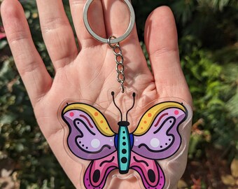 May include: A clear acrylic keychain featuring a colorful butterfly with yellow, pink, purple, blue, and white accents. The butterfly has a detailed design with dots and swirls.