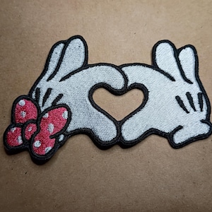 May include: Embroidered patch featuring white cartoon hands forming a heart shape. The patch includes a red bow with white polka dots on the left side and is outlined in black. The design is on a brown surface.