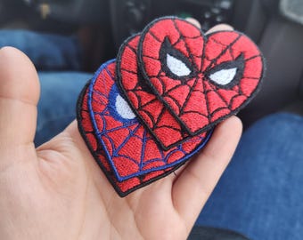 May include: Heart-shaped embroidered patches featuring the Spider-Man mask design. The patches are red with black webbing, white eyes, and a black border. One patch is blue with red webbing.