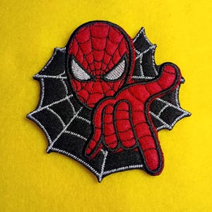 May include: Embroidered patch featuring a red Spider-Man design with a black spiderweb background. The patch has white embroidered eyes and a red body. The patch is set against a yellow background.