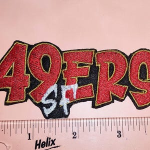 May include: Embroidered patch with the text "49ERS" in red, outlined in black and gold. The letters "SF" are in white. The patch is on a pink background. A ruler is at the bottom.